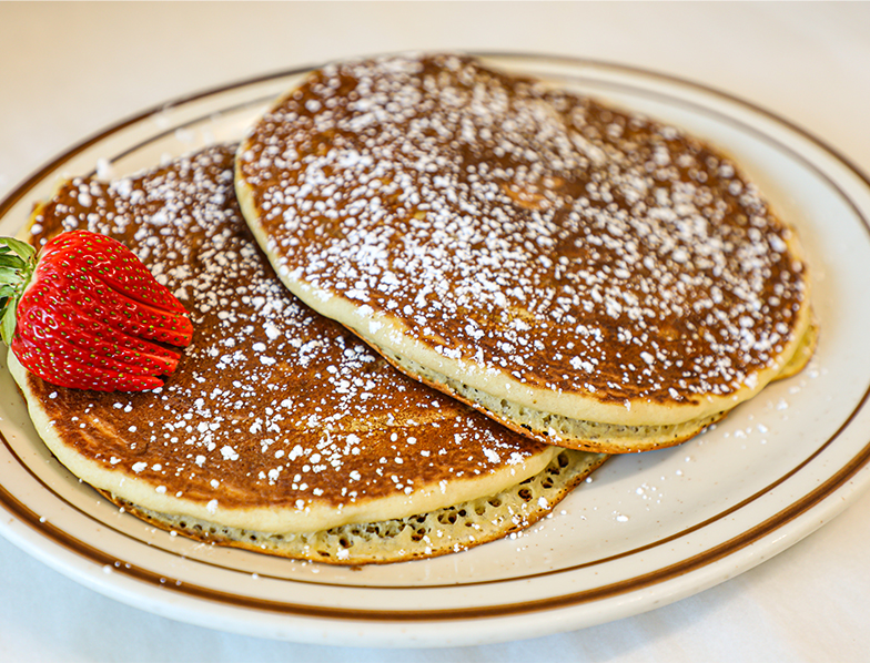 Two pancakes topped with powdered sugar and served with a fresh strawberry on a white plate with a brown rim.