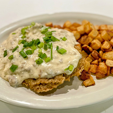 A plate with a serving of country-fried steak topped with gravy and chopped green onions, accompanied by a side of home fries.