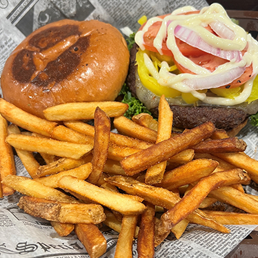 Burger with lettuce, tomato, onions, and yellow peppers on a bun, served with French fries on a newspaper-style liner.